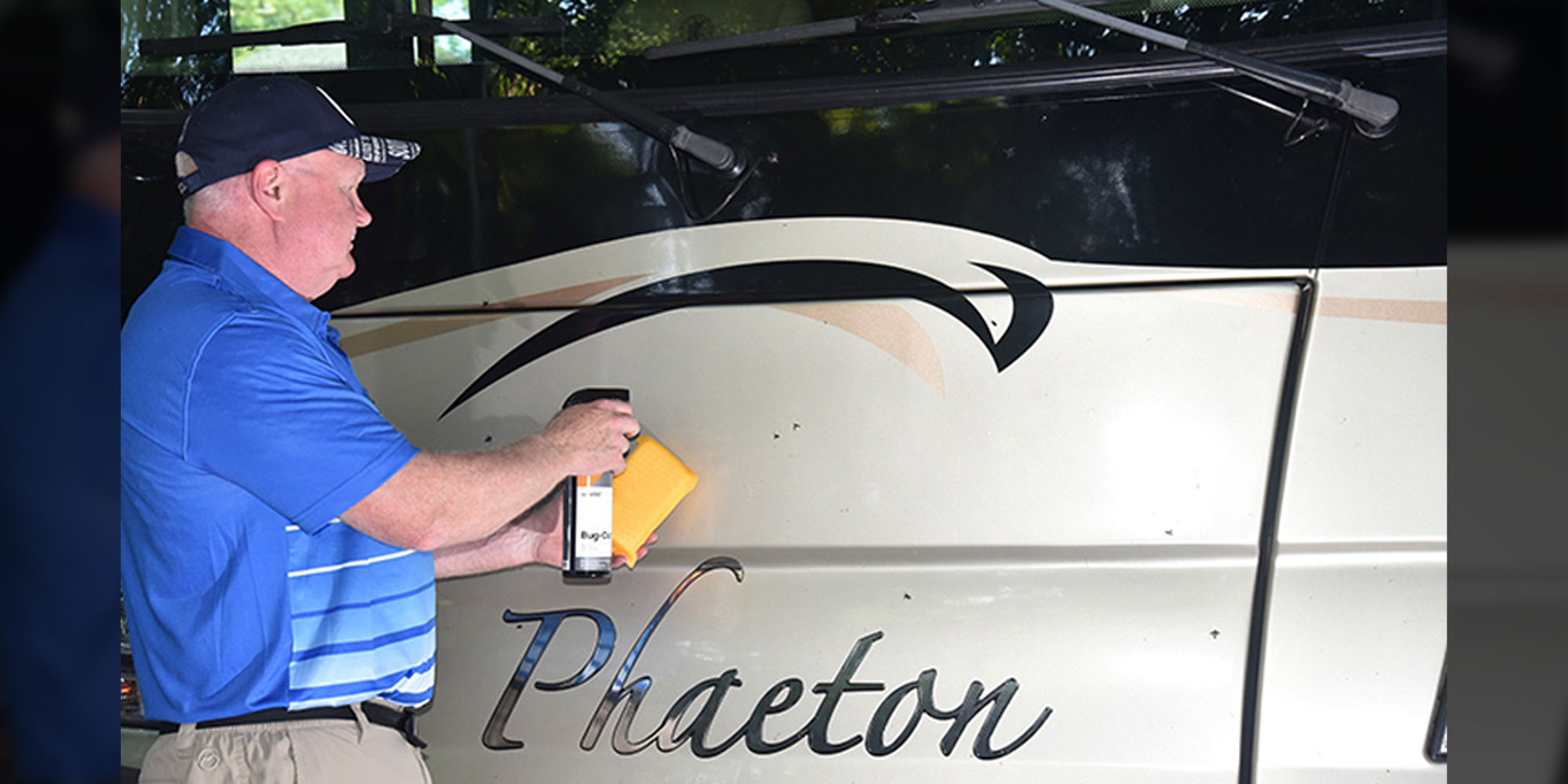 a man in a blue polo shirt applies a cleaner to the front of an RV while holding a sponge in his other hand