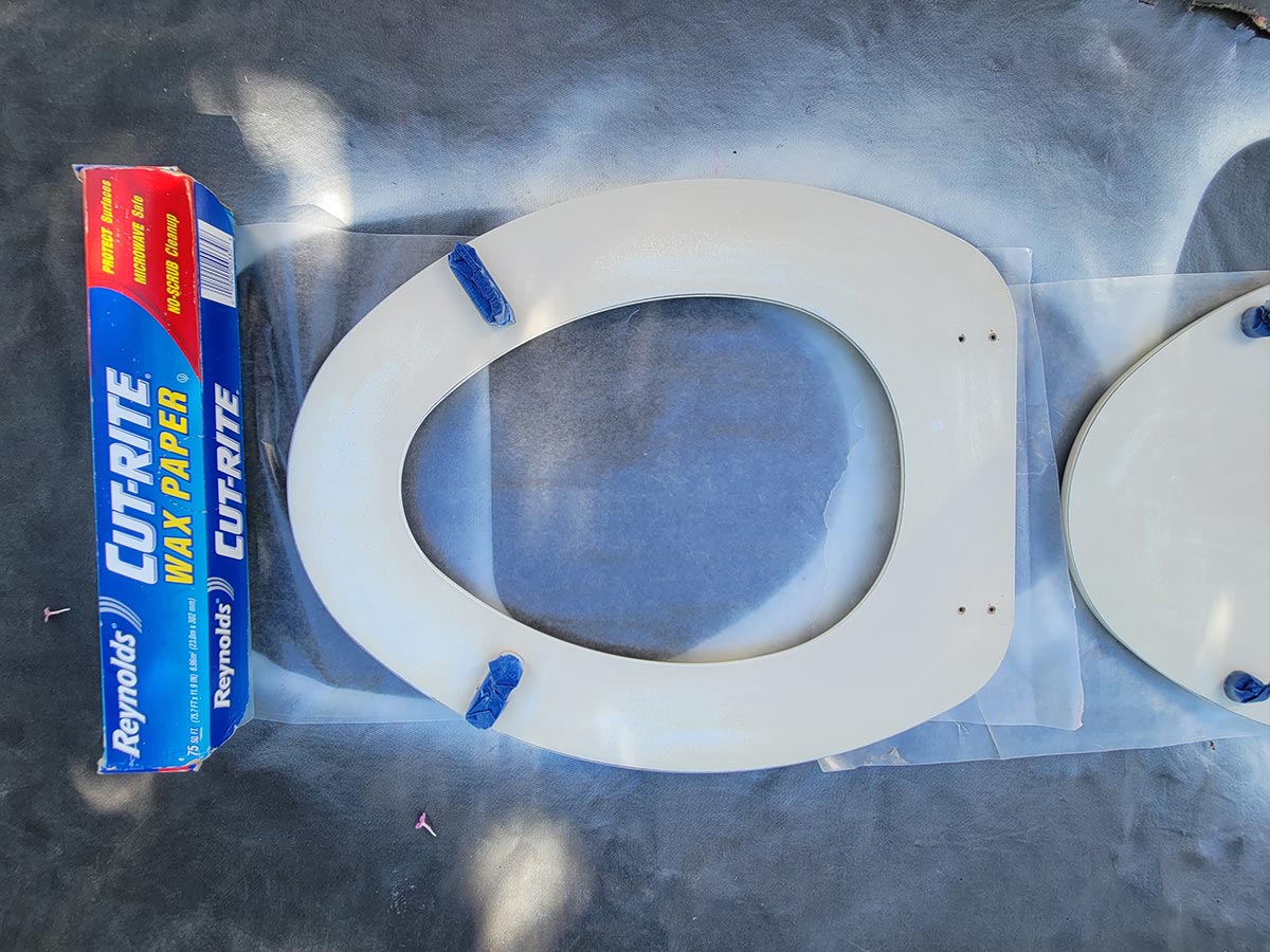 the freshly painting toilet seat is placed on wax paper to protect the paint from sticking