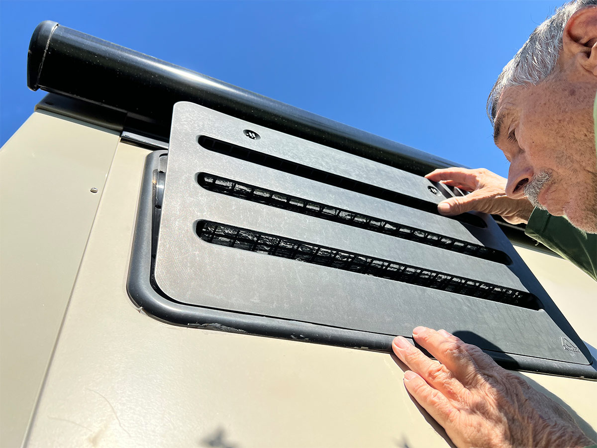 RV technician removes a vent covering installed in the sidewall behind the absorption refrigerator