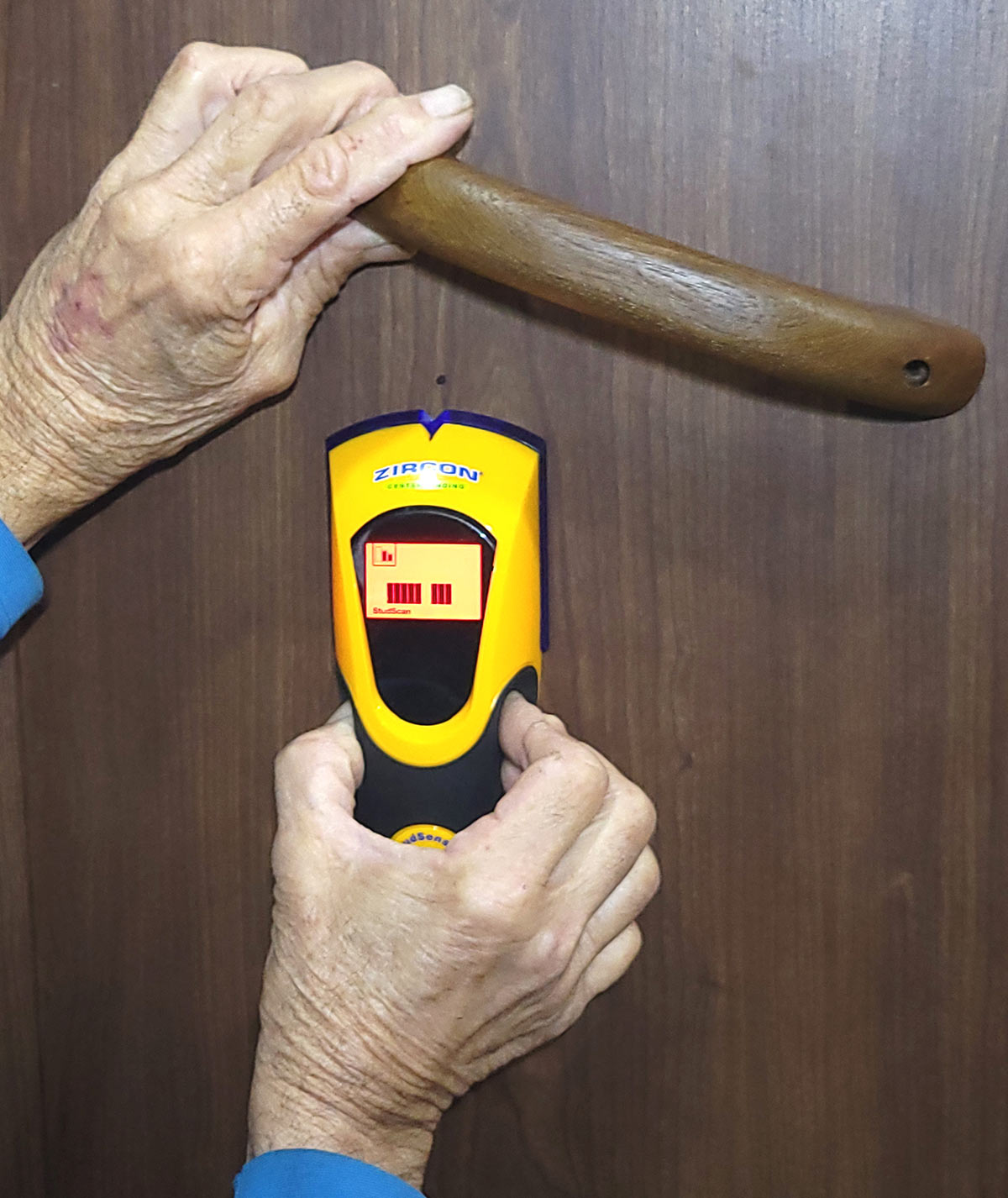 hands use a stud finder to find the stud behind a wood panel holding the current wooden rail
