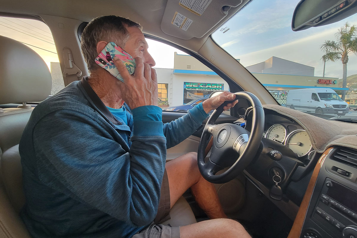a man sits at the driver seat of a vehicle with one hand on the wheel and another holding a phone to his hear
