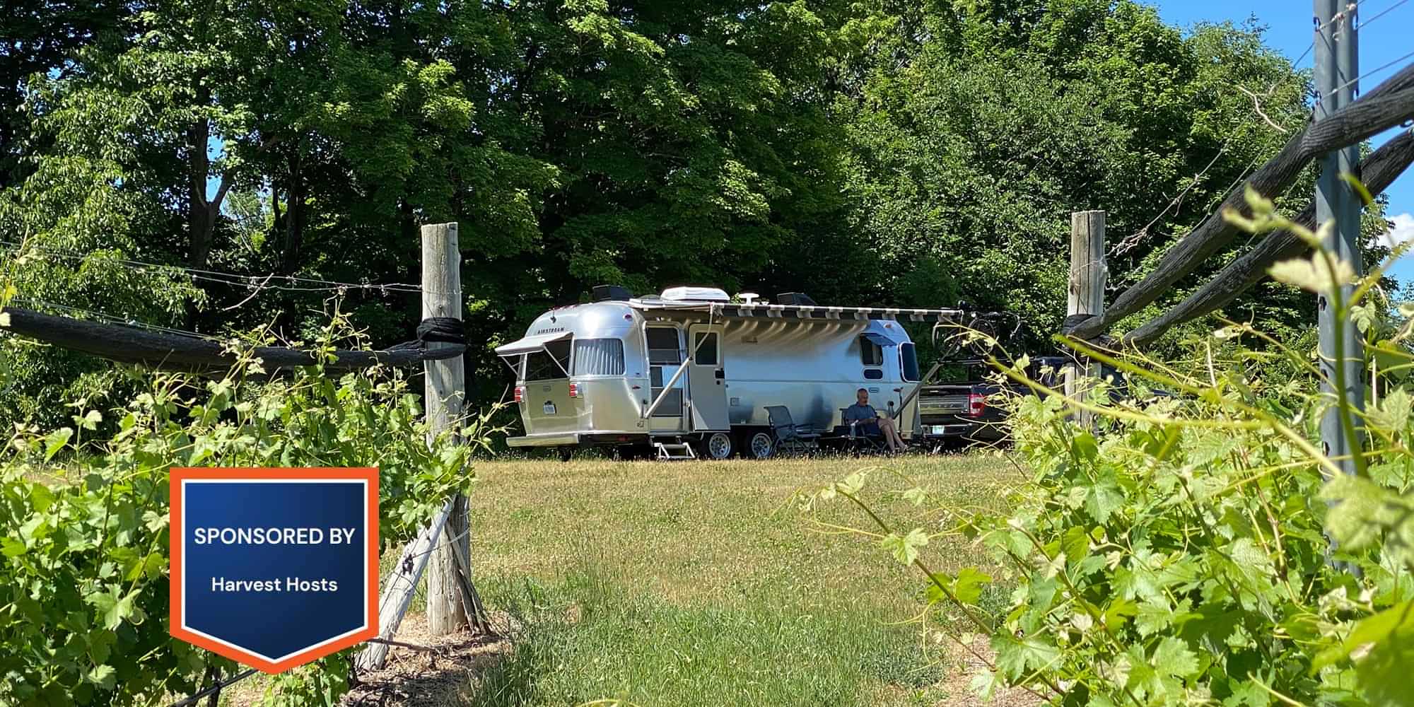 a man relaxes on one of two lawn chairs under the awning of his vintage looking travel trailer parked among trees and shrubs
