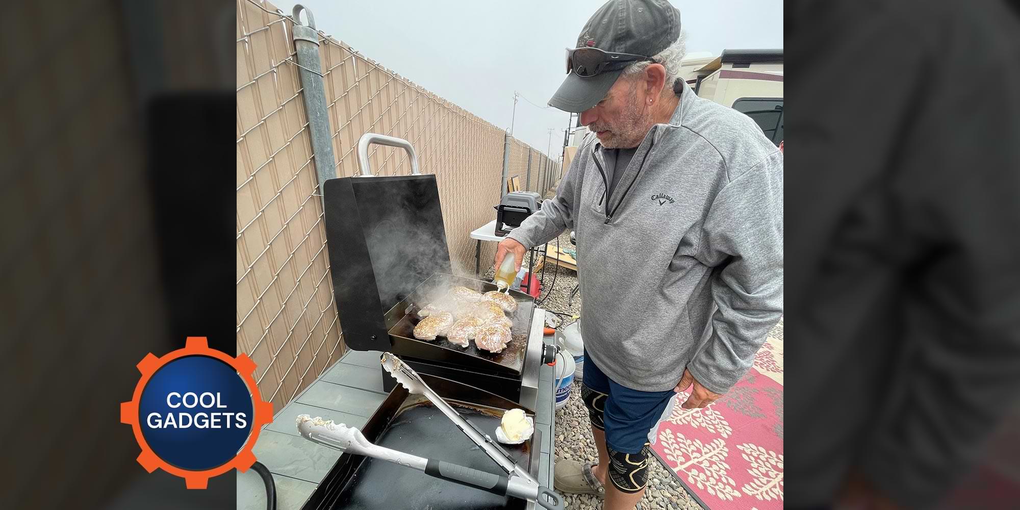 a man stands cooking with a table top grill near a fence and RV