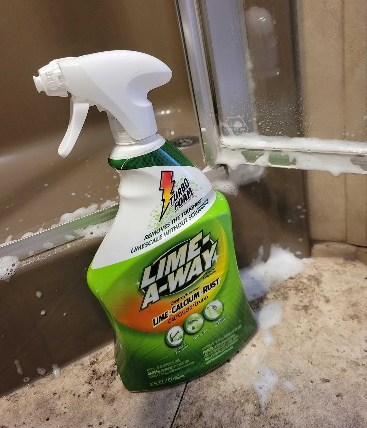 a Lime-A-Way container placed on the floor in front of a shower door bottom that has bee sprayed