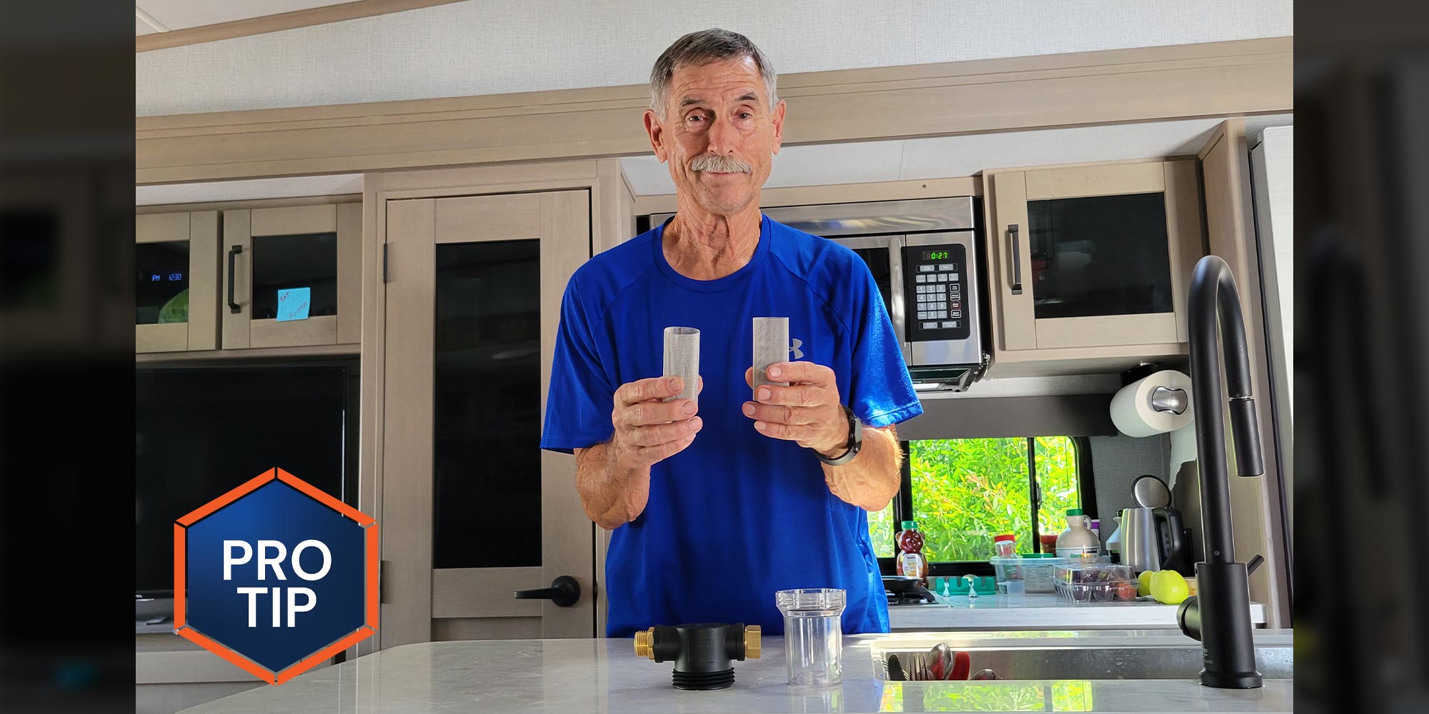 a man in a blue shirt stands at an RV kitchen counter while holding a mesh cylindrical filter in either hand, a device and a small plastic container sit on the counter in front of him