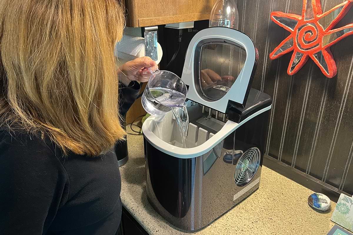 a woman pours water from a measuring cup into a portable ice maker on the counter of an RV kitchen
