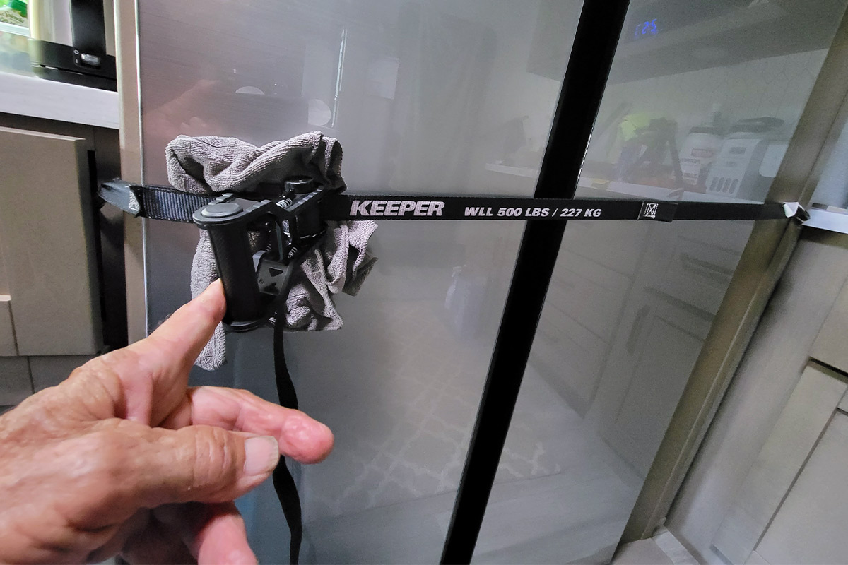 a finger points to a ratcheting strap with hooks on the ends being used to keep an RV's refrigerator doors closed