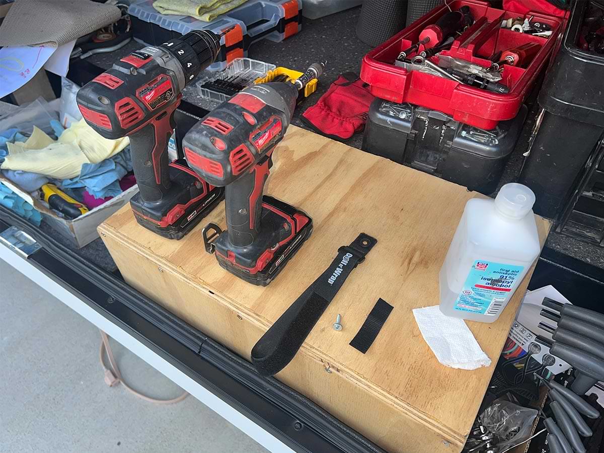 two drills, an old Coil n’ Wrap nylon strap, and a container of Isopropyl Alcohol sit a on wood box at the opening of an RV tool storage compartment