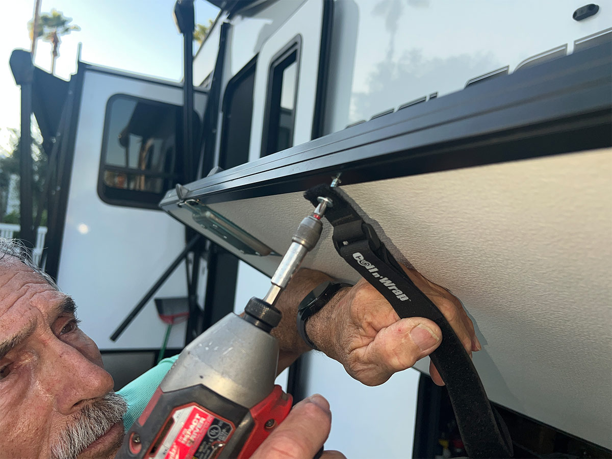 the RV technician carefully drills the lath screw through the strap grommet into the compartment door's edge molding