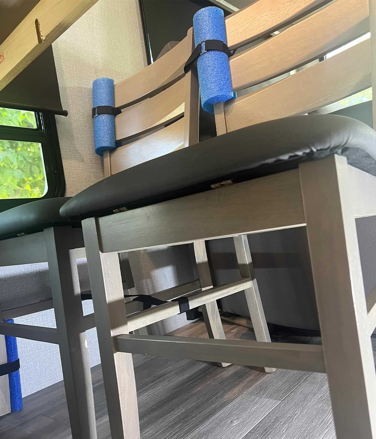 two RV dinette chairs with blue swim noodle pieces protecting the areas where they touch are also secured to the RV wall by a single strap fastened to the Footman Loop