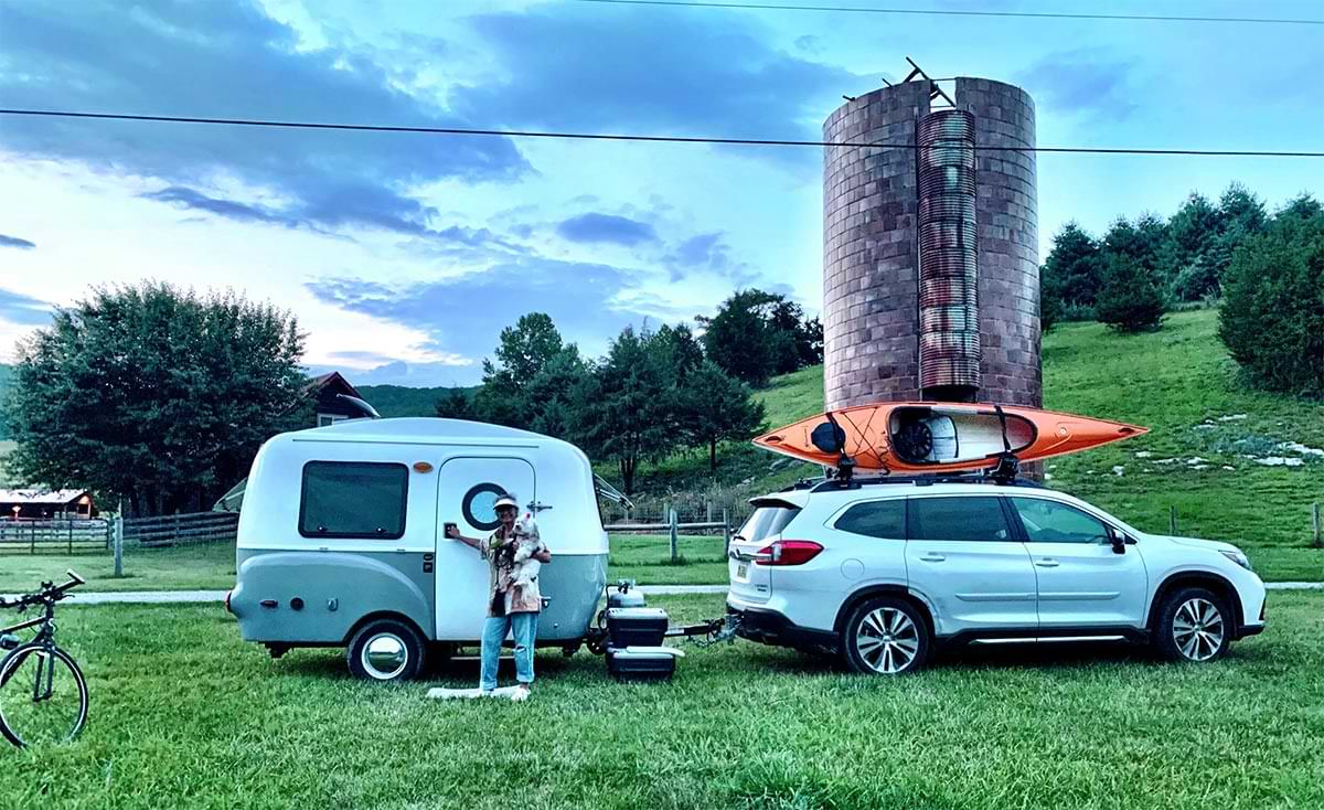 a woman stands at the door of a small camper in a rural field beside a hill covered with trees, the camper is hitched to an SUV with a kayak on top and the woman smiles while holding a small breed dog