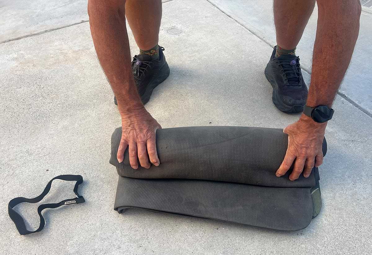 using the ground as a surface, hands tightly roll a folded mat as a strap lays near by