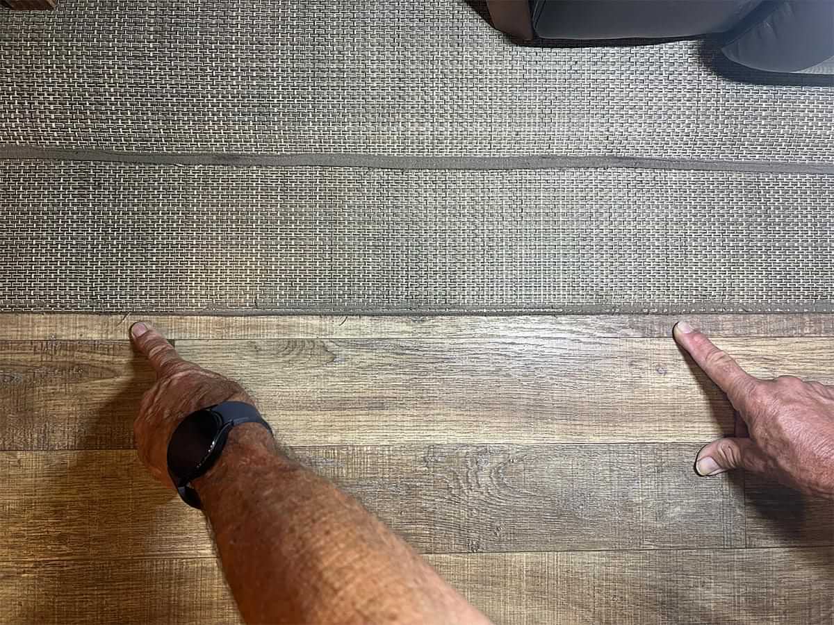 two index fingers point to areas of tattered binding along the edge of an RVs woven vinyl flooring