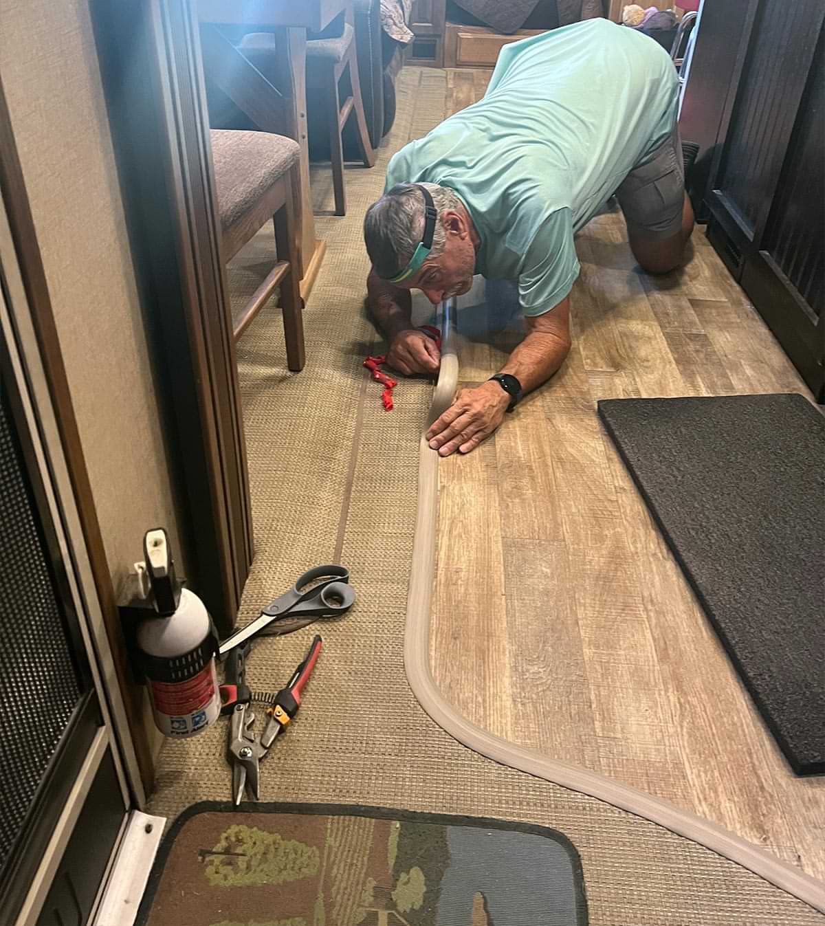 a man wearing a headlight leans low to the floor while carefully removing tape backing and adhering the transition strip along the edge of woven vinyl flooring