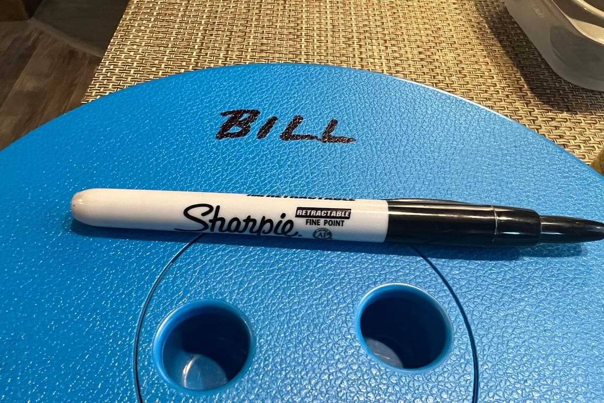 close view of a blue plastic telescoping stool with a Sharpie resting on top, beside the sharpie reads the word: BILL, written on the stool seat