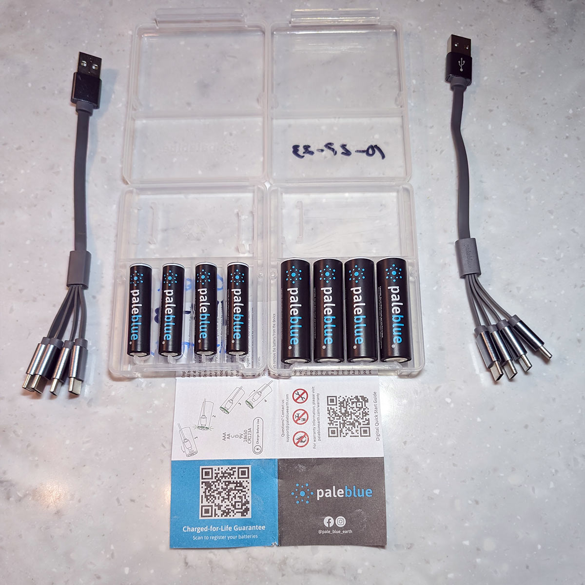 top view of an open plastic container holding four triple A Paleblue batteries and four double A Paleblue batteries along with two USB cords, each with four USB-C adapted ends, and instructions