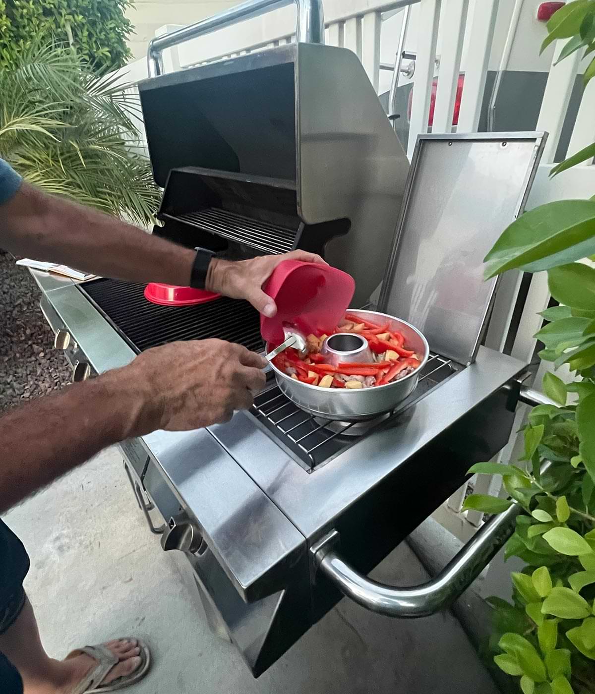hands pour food ingredients into the molded Omnia pan that sits on a side grate of a large grill