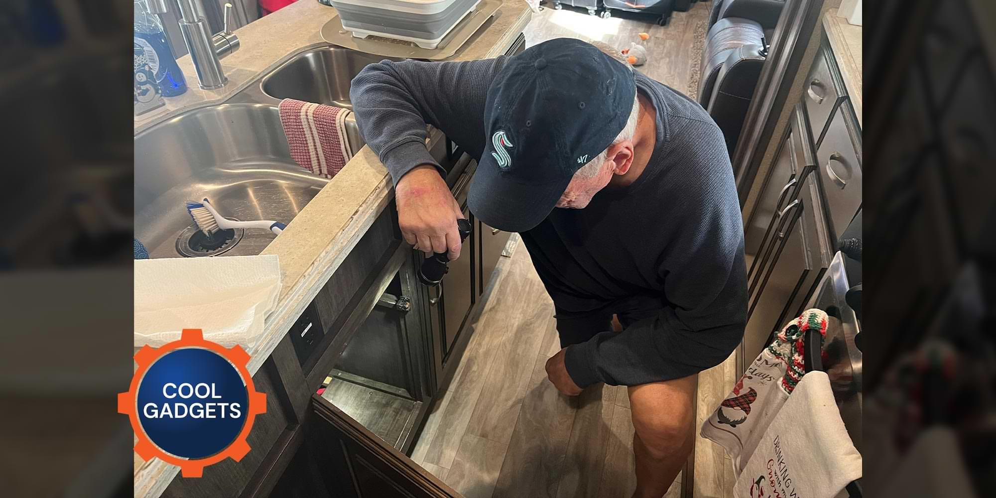a man holding a flashlight kneels looking into the cabinet beneath an RV sink