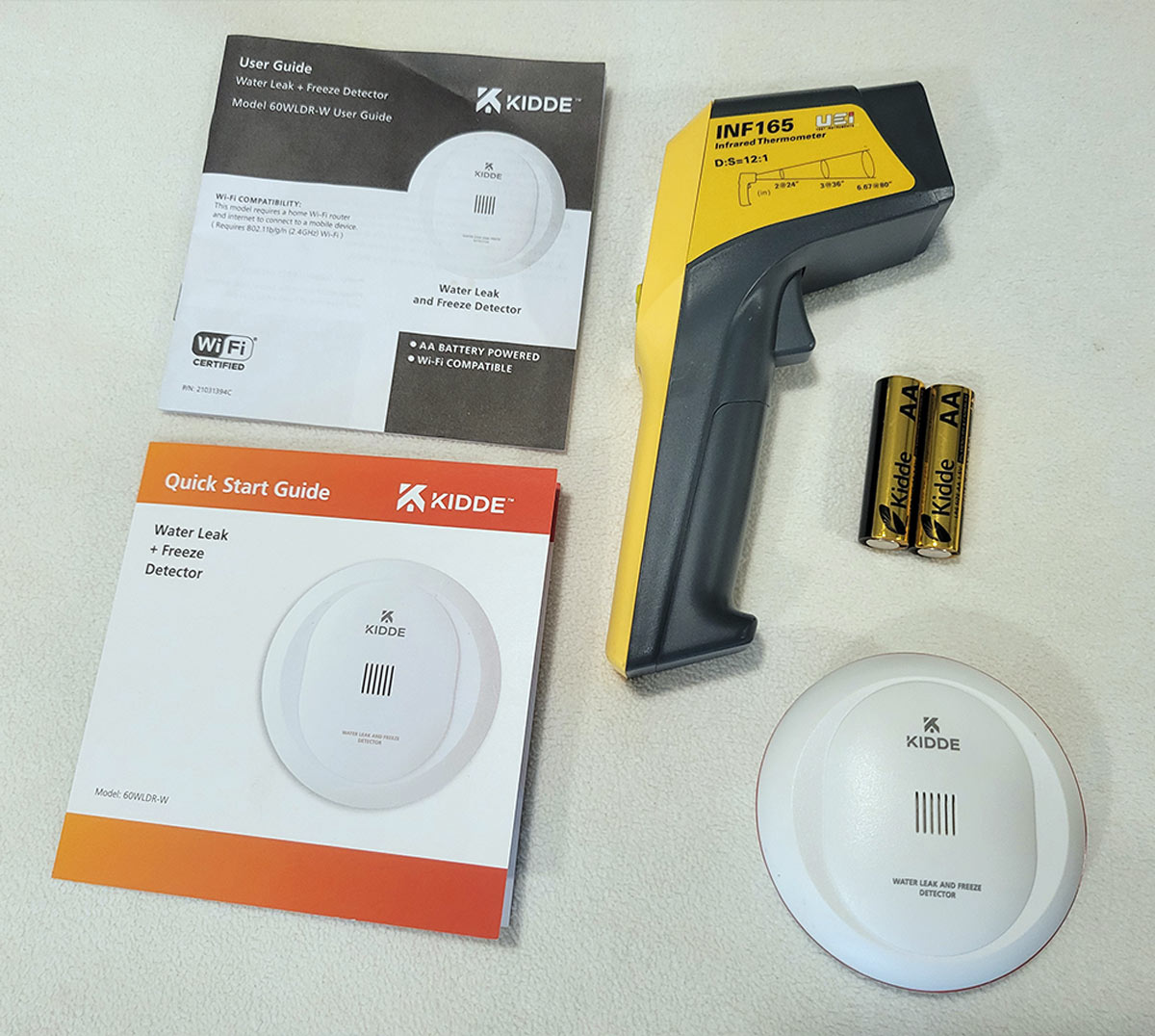 top view of a temperature gun, the Kidde freeze and water detector, the detector direction and two double A batteries 