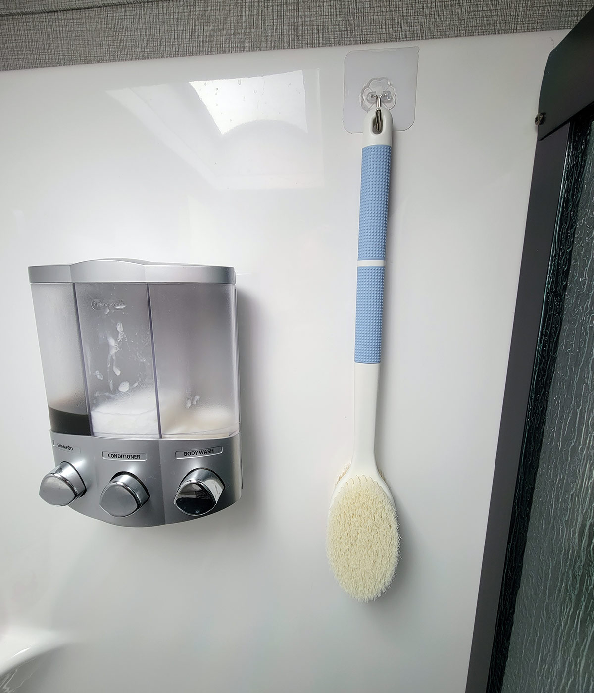 close view of a body brush hanging from a JelKen transparent base Wall Hook beside a body wash, shampoo and conditioner dispenser in an RV bathroom