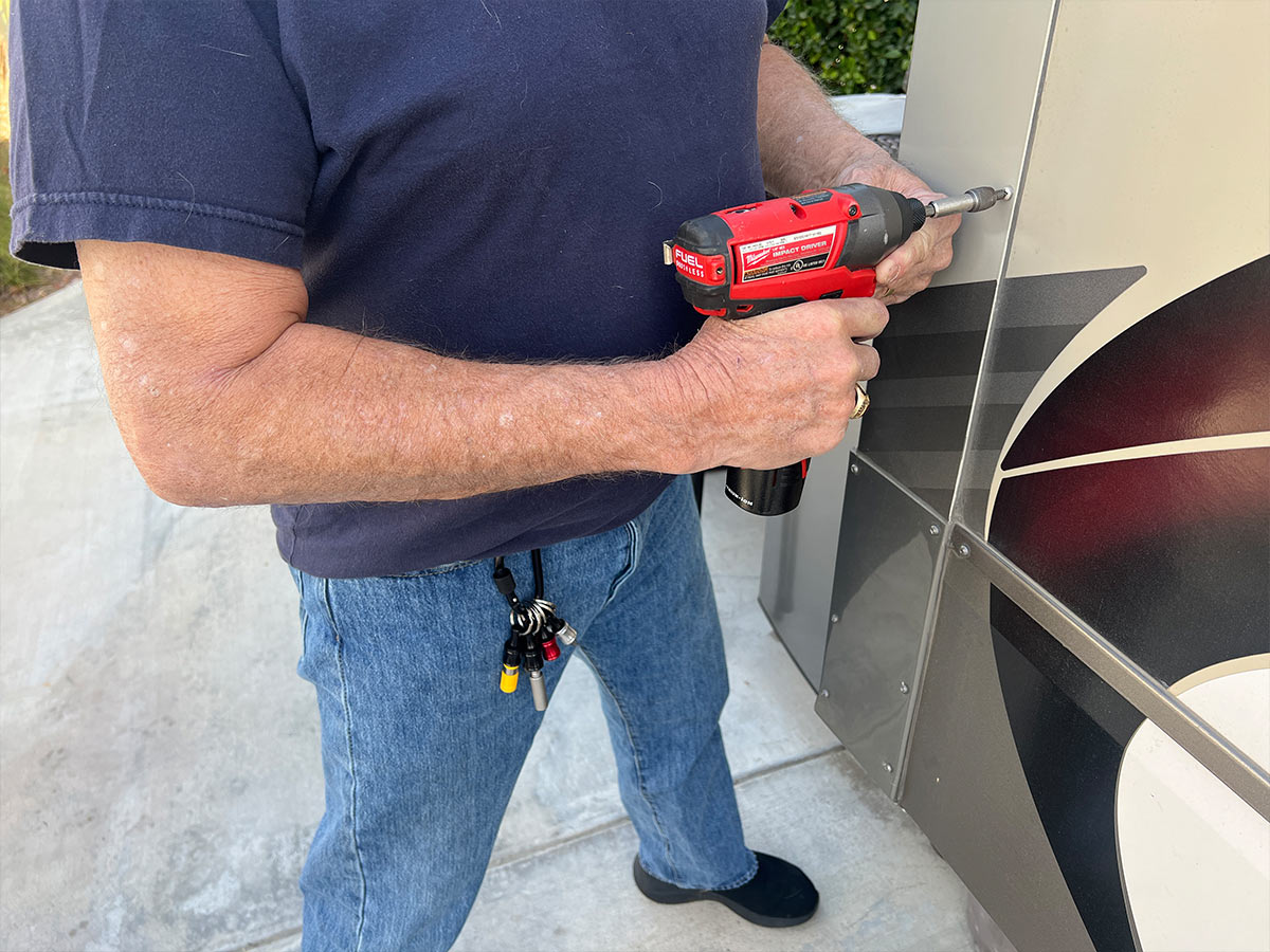 cropped view of a man holding a drill gun to a screw on the side of an RV, on a front belt-loop of the man's jeans hangs a carabiner with a group of bit holders