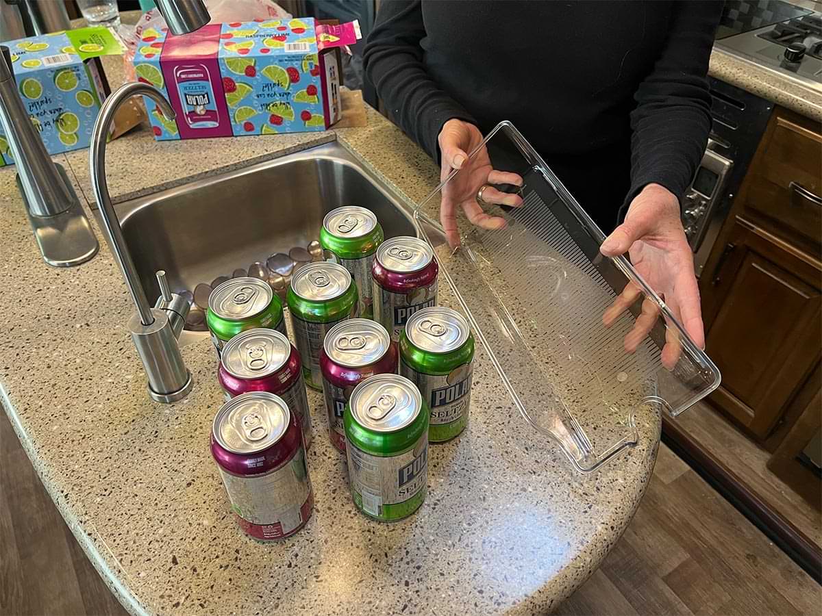 unpacked cans sit on an RV kitchen counter top, beside the cans a woman holds a dispenser tray