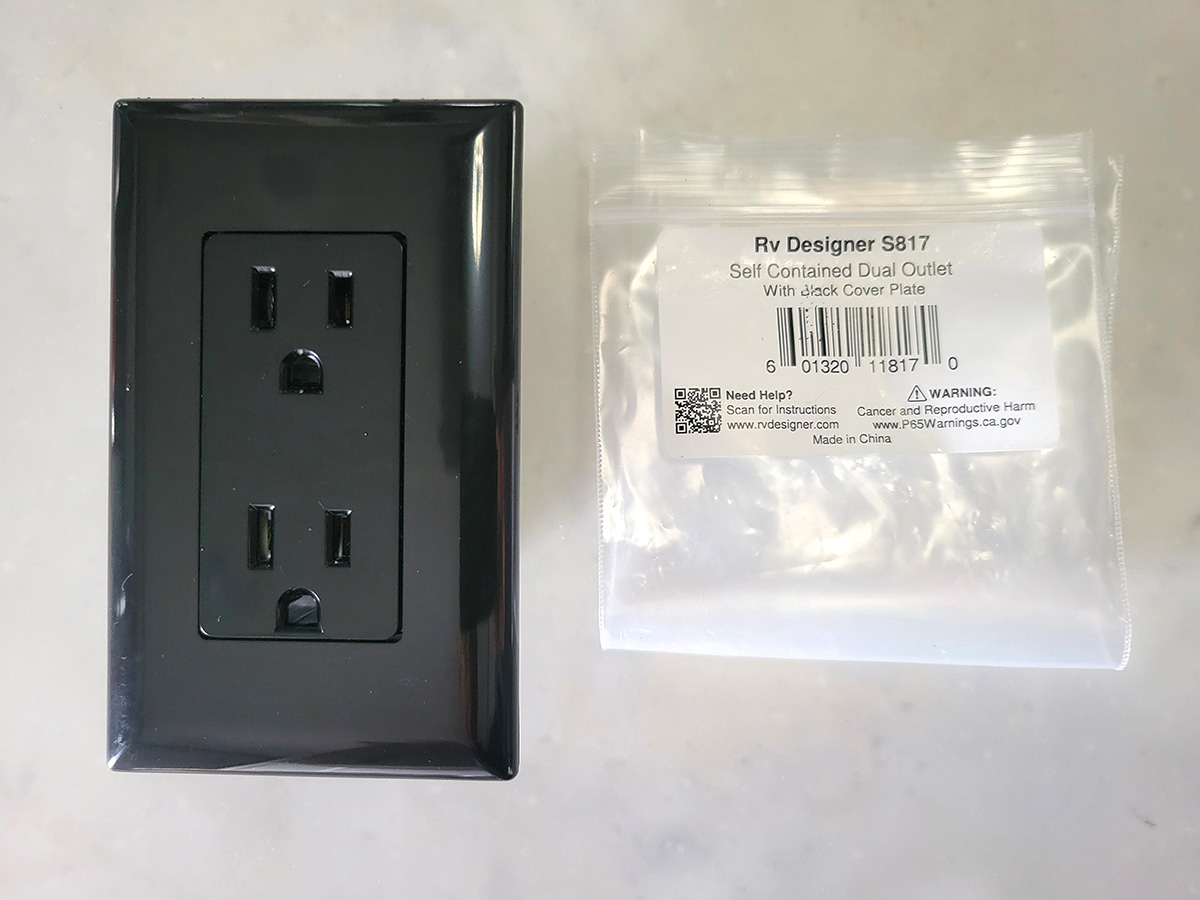 close view of a black one-piece RV-type 120-volt AC outlet beside its package bag