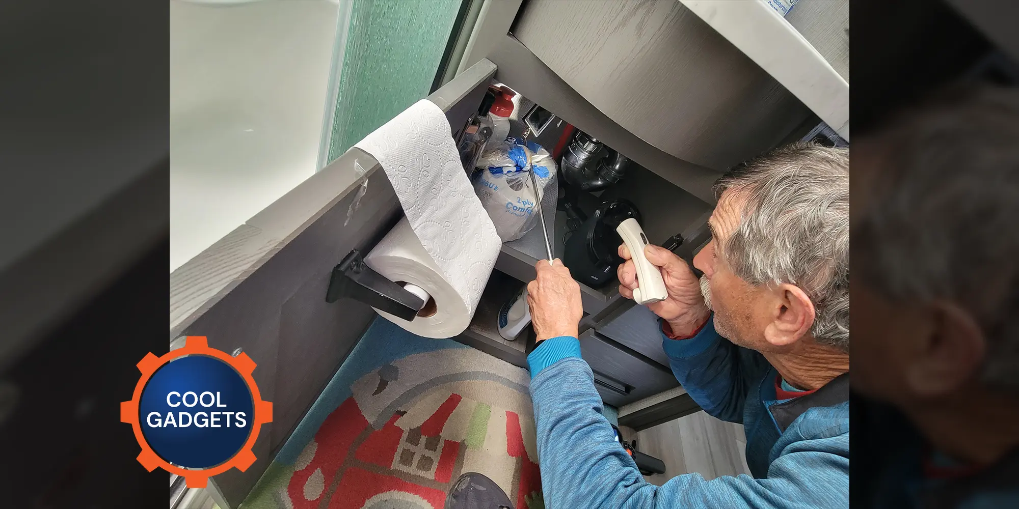 a man uses a small flashlight and extendable mirror to look at piping in a small undersink compartment in an RV