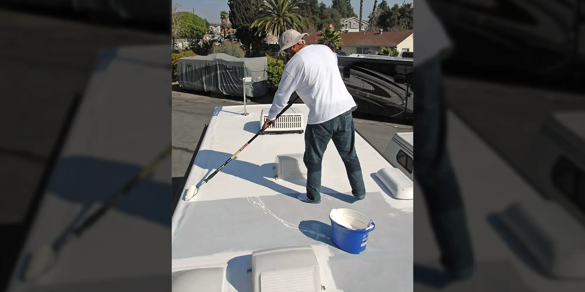 a man atop an RV, using a paint roller to apply a coat of a rubber sealant