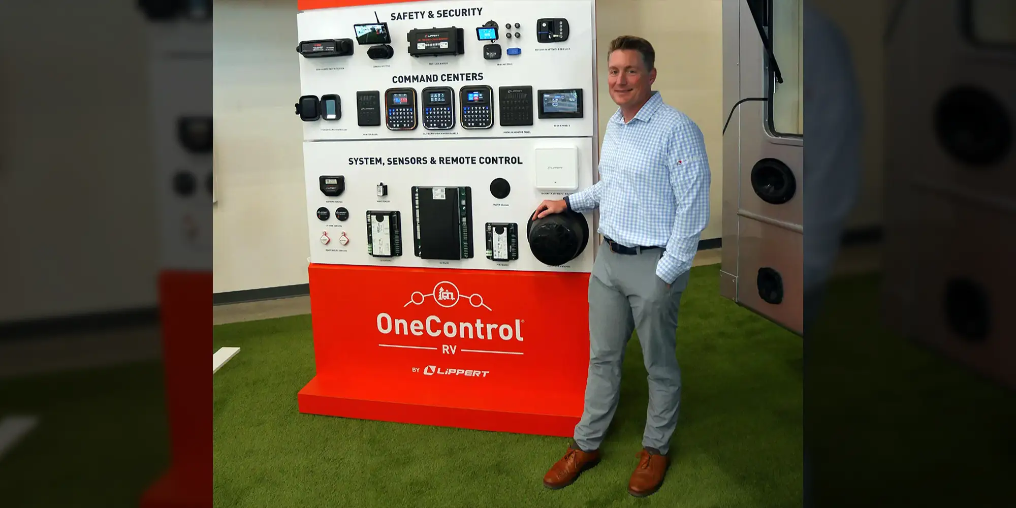 Jarod Lippert wearing a light blue plaid button up shirt and slacks smiles while standing alongside a OneControl display showing the wealth of functions the device now controls