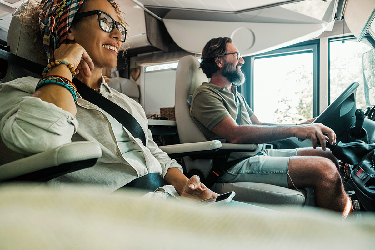 low angle view from the passenger side of a man and a woman sitting in the driver and passenger seat of an RV both smiling while looking at the road