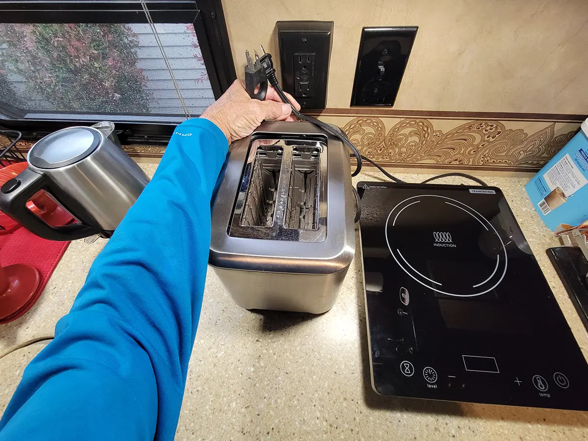 wide view of an RV galley counter top holding an induction hot plate and a toaster, a hand holds the plugs for both appliances near a galley wall outlet