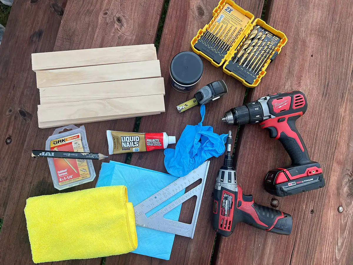 the tools for the shelf bracing project laid on a picnic table including, eight wooden 1 x 2’s, wood stain, screws, drill guns and bits, a set square, measuring tape, a pencil and a microfiber towel