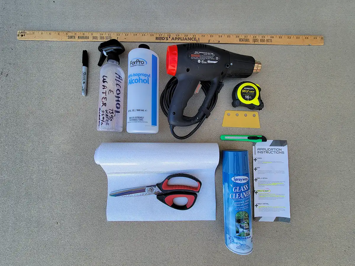 top view of the tools for the window film installation, including a ruler, a sharpie marker, a spray bottle, Isopropyl alcohol, a heat gun, a tape measure, a plastic squeegee, scissors, a razor knife, vinyl film and glass cleaner